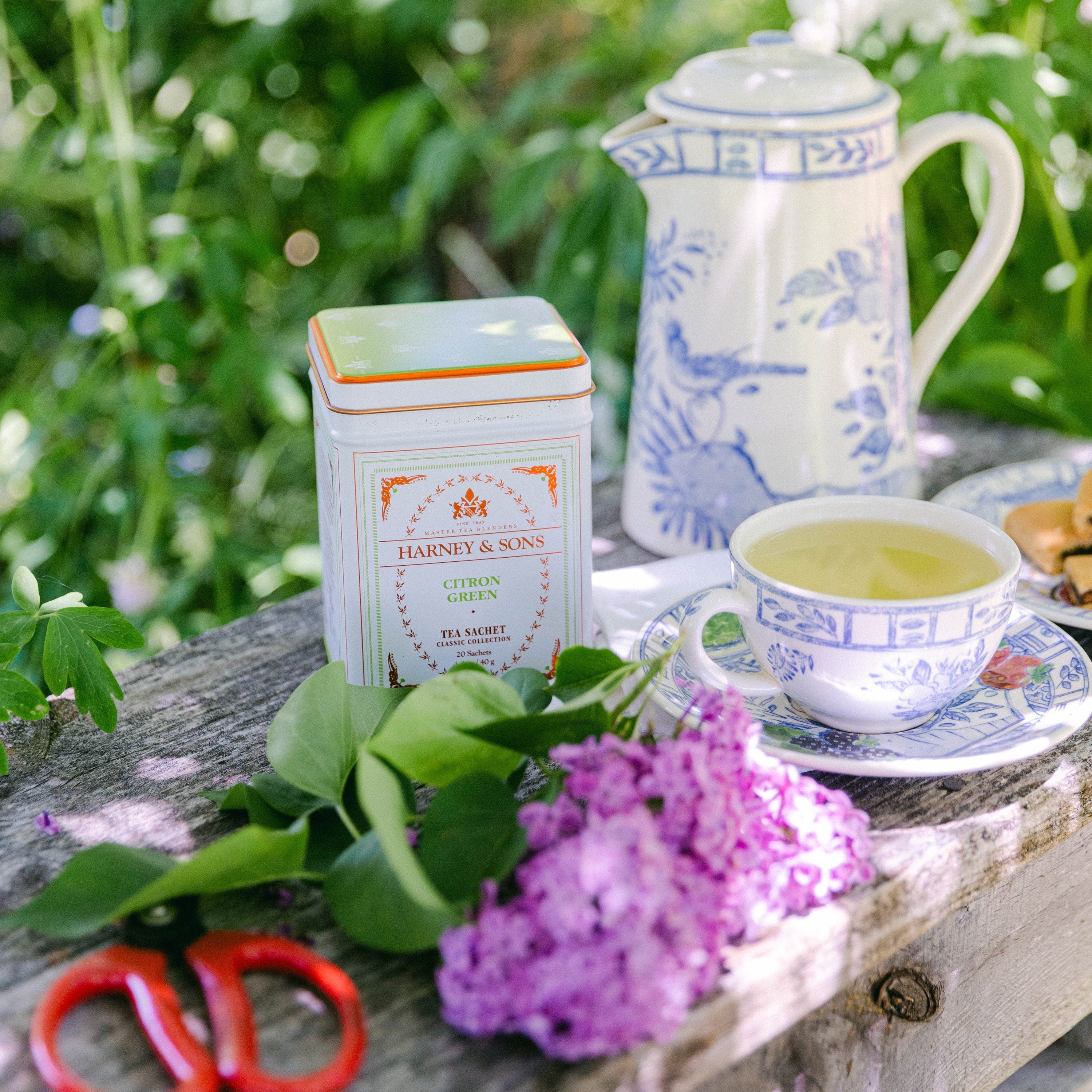 A tea setup on a rustic wooden table outdoors, featuring a white and blue porcelain teapot, a cup of tea on a saucer, a white tea tin labeled Harney & Sons, green leaves, purple flowers, and red scissors, with a blurred garden background.