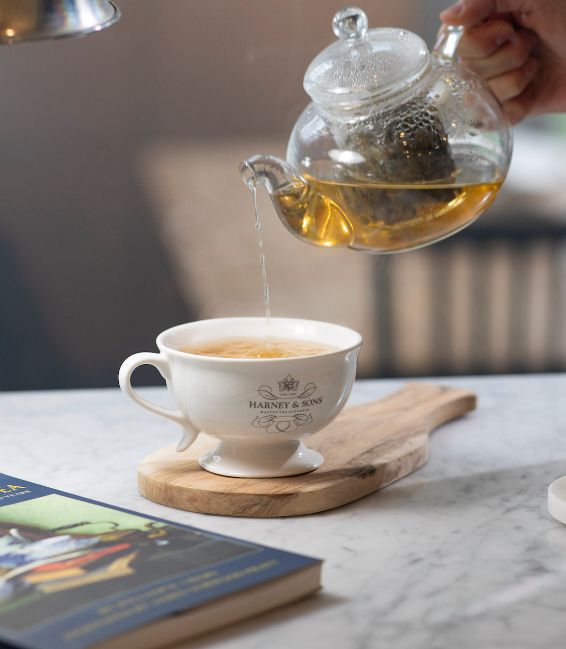 Glass teapot pouring tea into a white cup on a marble surface with a blurred background.