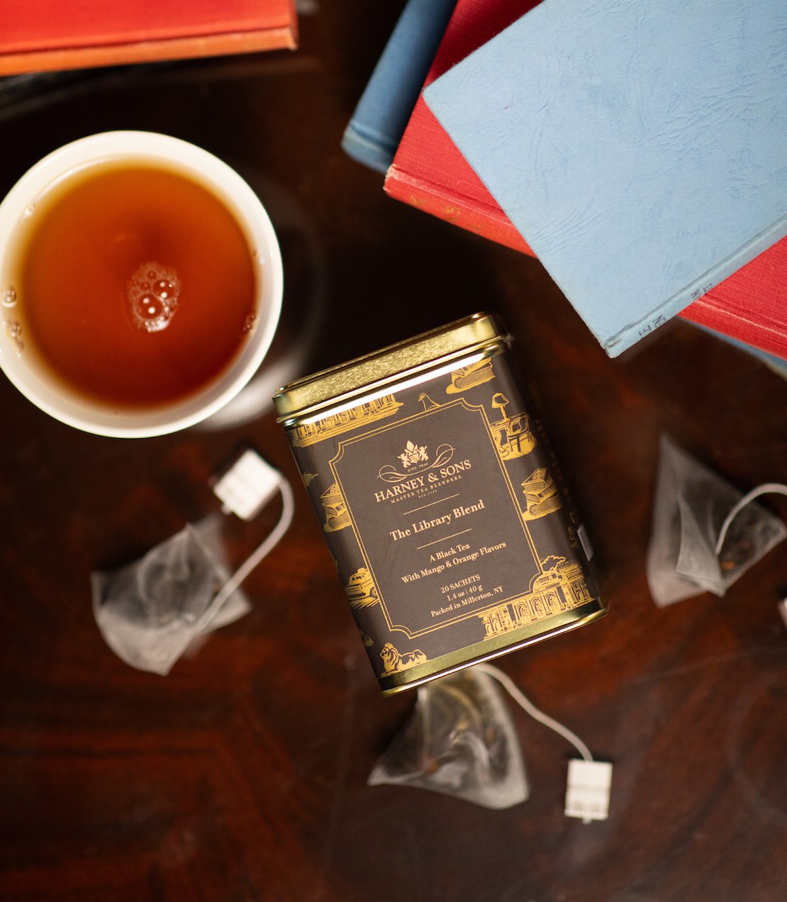 Harney & Sons Fine Teas- The Library Blend Tin of 20 sachets- Gold tin with black and gold label- Pictured from above, surrounded by tea sachets, a cup of tea, and books. 